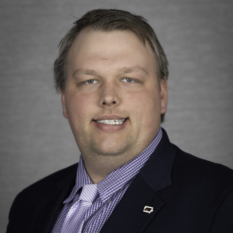 Professional portrait of a man in a navy blazer and purple checkered shirt with a tie, smiling against a gray background.