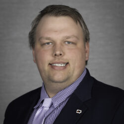 Professional portrait of a man in a navy blazer and purple checkered shirt with a tie, smiling against a gray background.